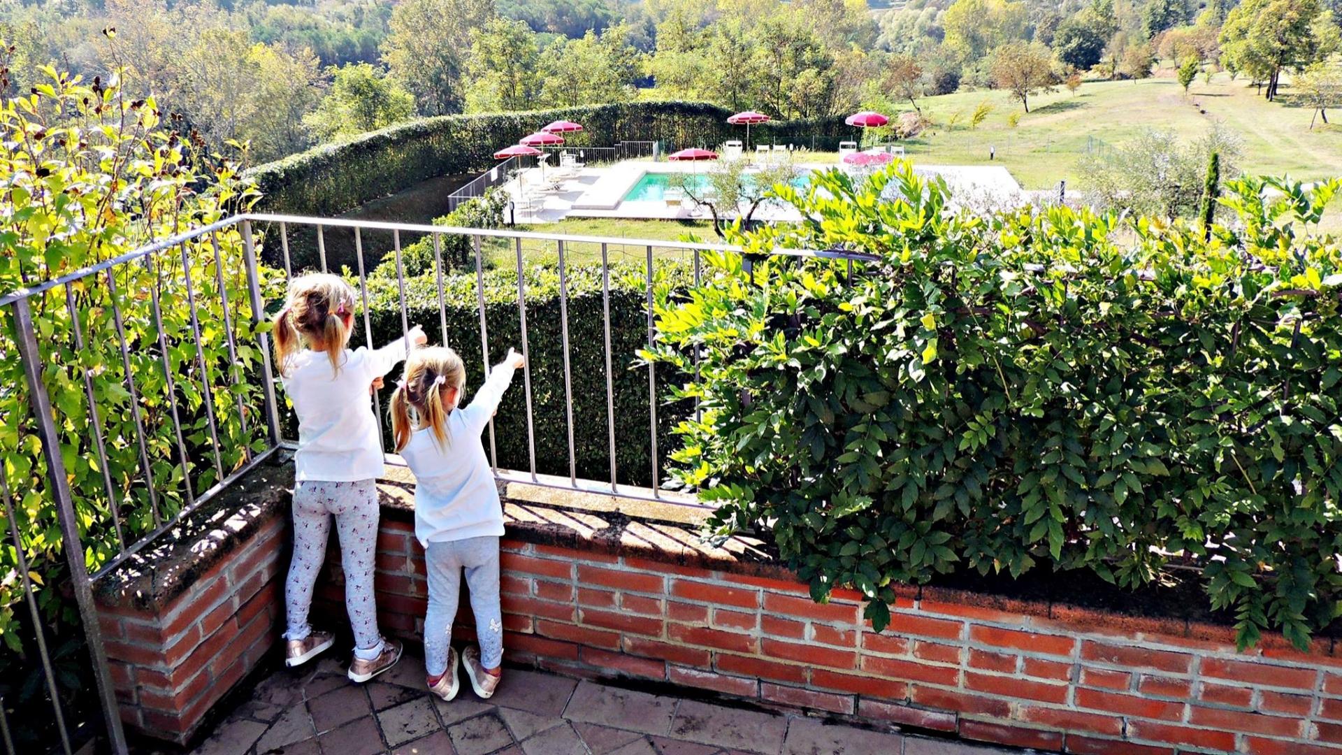 Due bambine guardano una piscina circondata da ombrelloni rossi in un paesaggio verde.