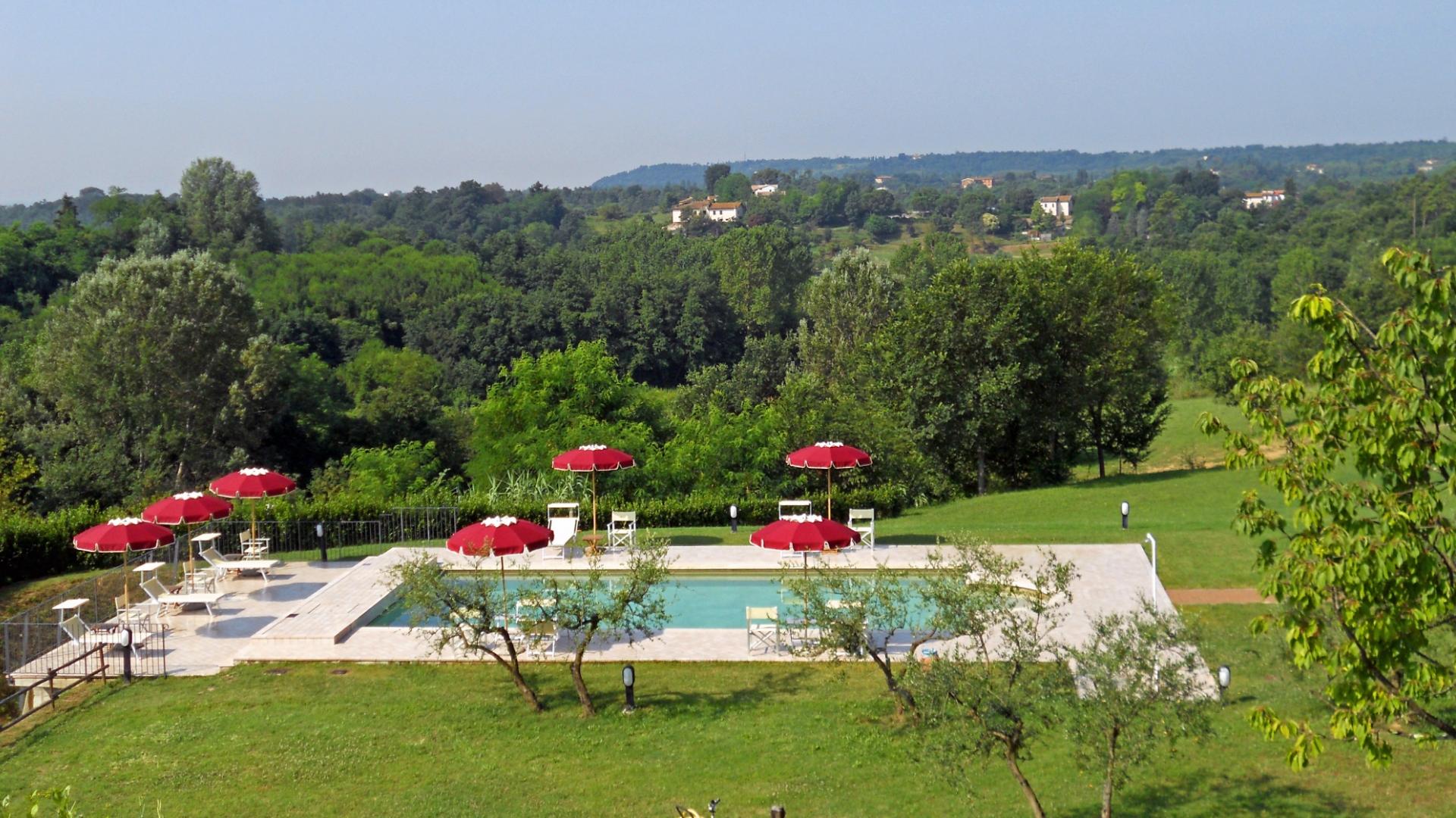 Piscina con ombrelloni rossi immersa nel verde paesaggio collinare.