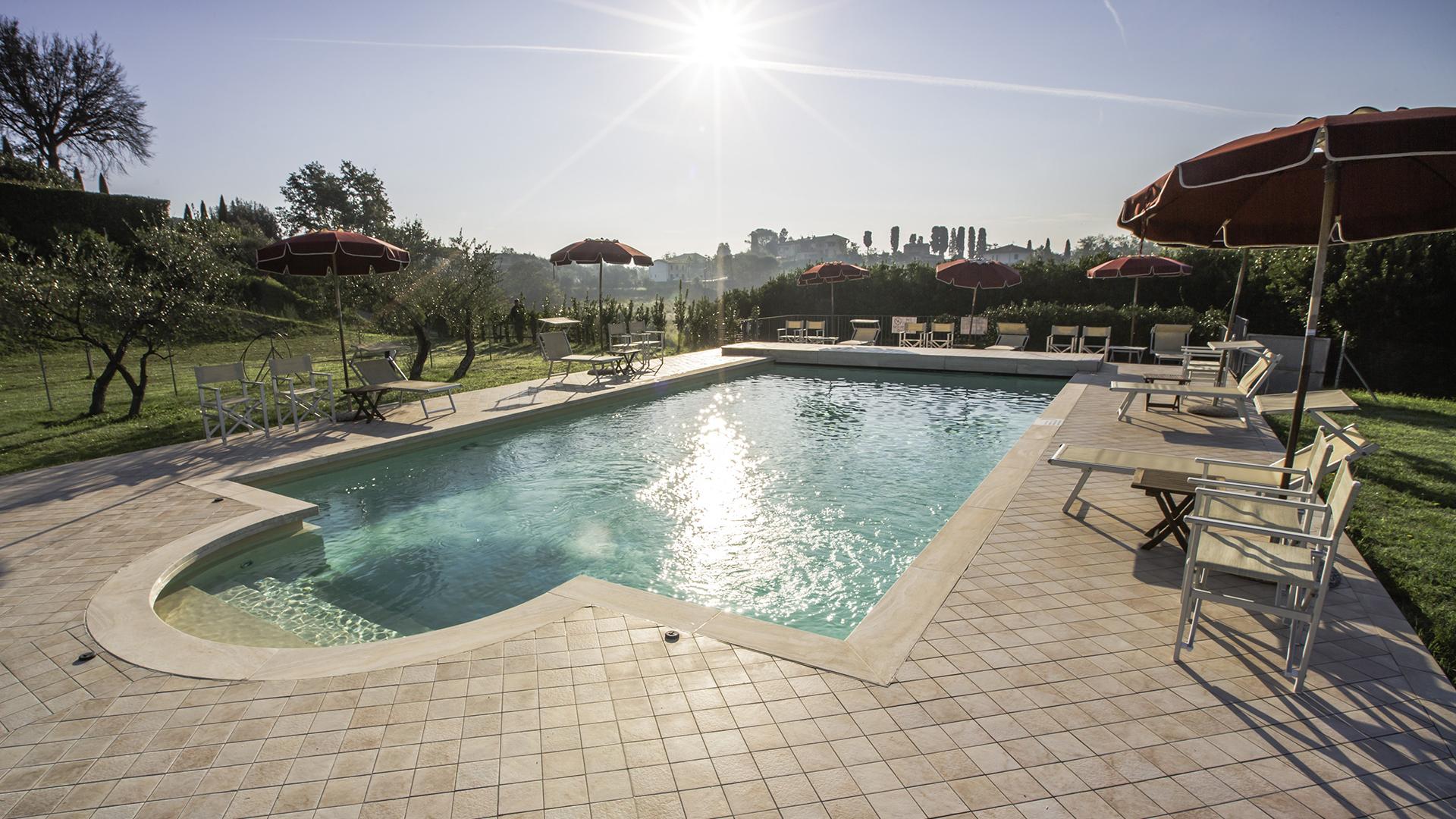 Piscina all'aperto con ombrelloni rossi, circondata da natura e sole splendente.