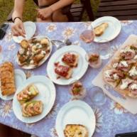Tavola estiva con bruschette assortite, pane e vino su tovaglia floreale.