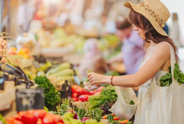 Donna al mercato sceglie verdure fresche, indossa cappello e borsa ecologica.