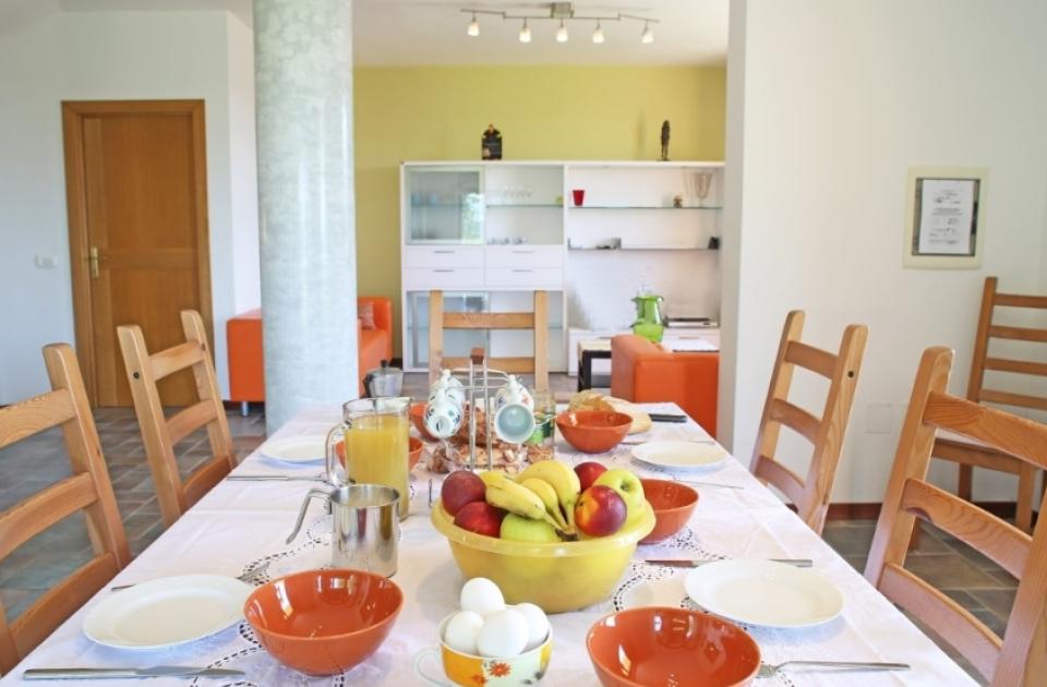 Tavola apparecchiata per colazione con frutta, uova, succo e ciotole su tovaglia bianca.