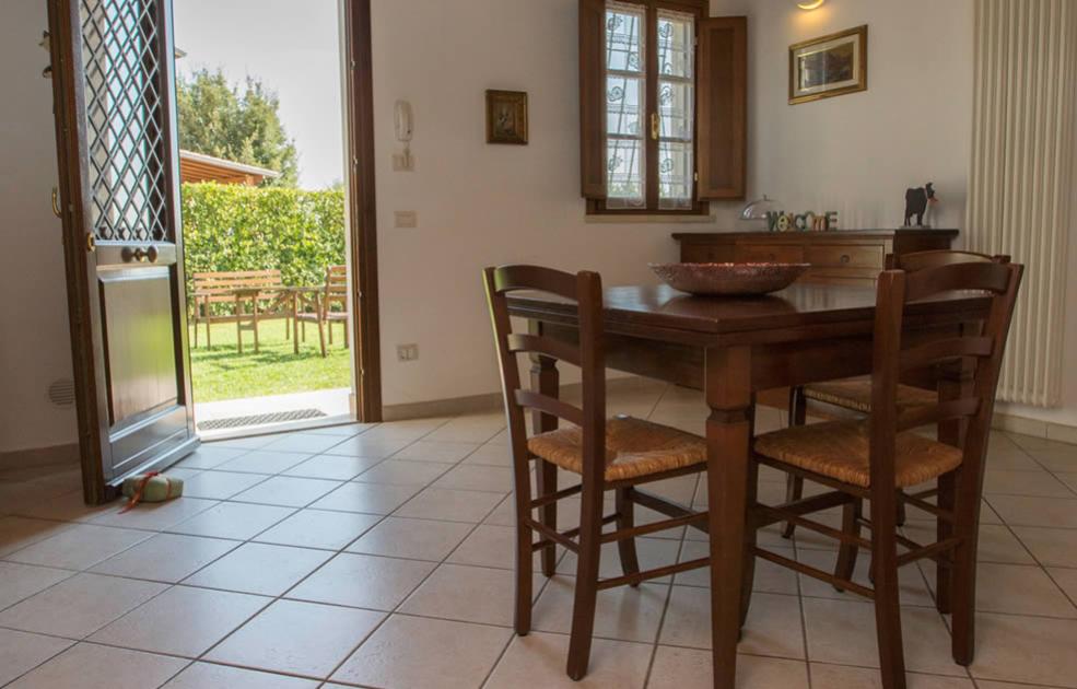 Sala da pranzo con tavolo in legno, porta aperta verso giardino soleggiato.