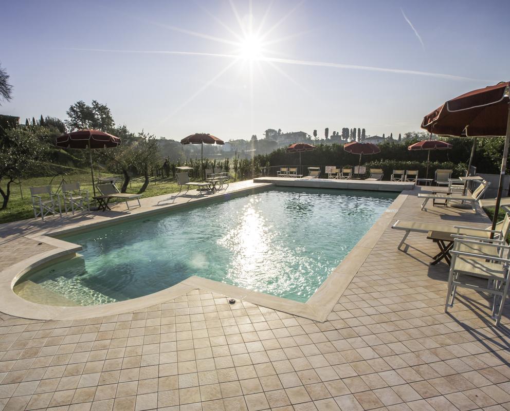Piscina all'aperto con ombrelloni rossi, circondata da natura e sole splendente.